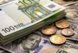 IMAGE: A close-up of stacked euro banknotes and scattered euro and US dollar coins resting on US dollar bills, symbolizing the persistence of physical cash in modern economies
