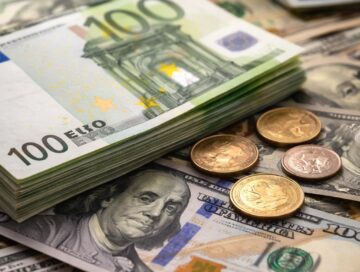 IMAGE: A close-up of stacked euro banknotes and scattered euro and US dollar coins resting on US dollar bills, symbolizing the persistence of physical cash in modern economies